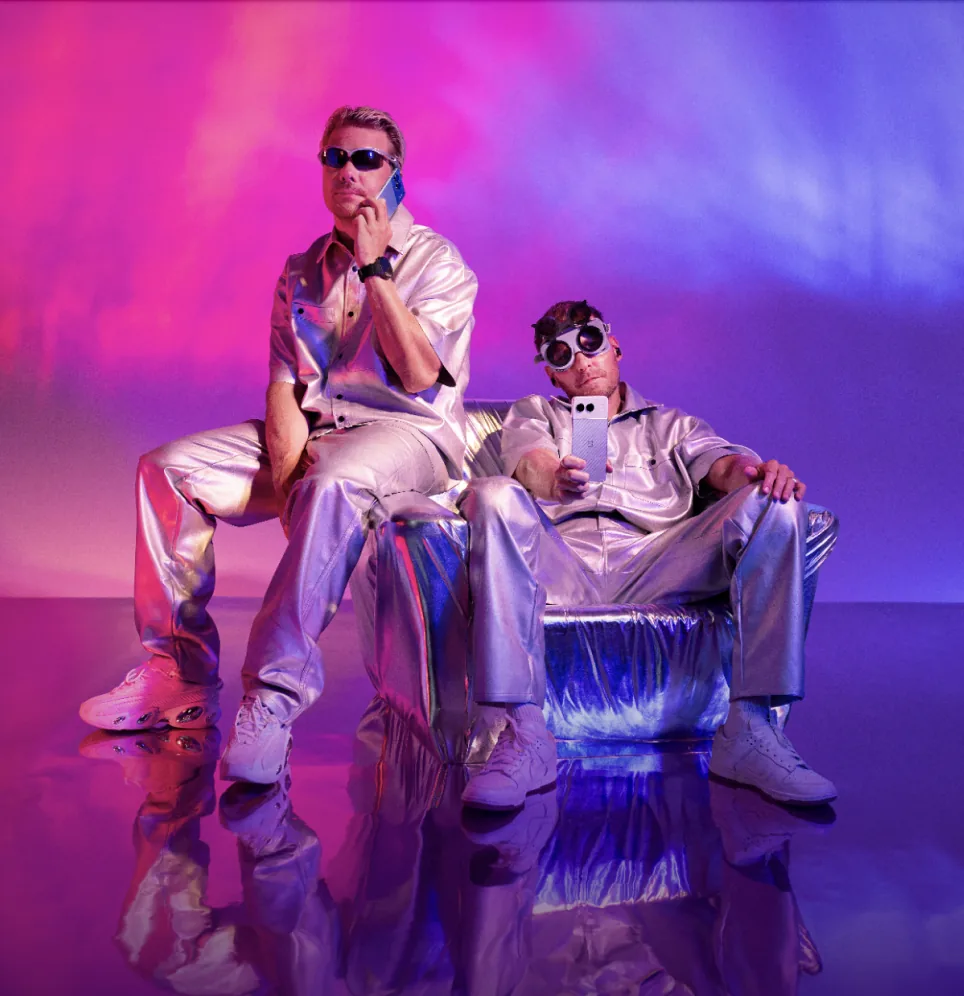JVG in silver suits on a metallic sofa, holding OnePlus phones, under pink and purple light